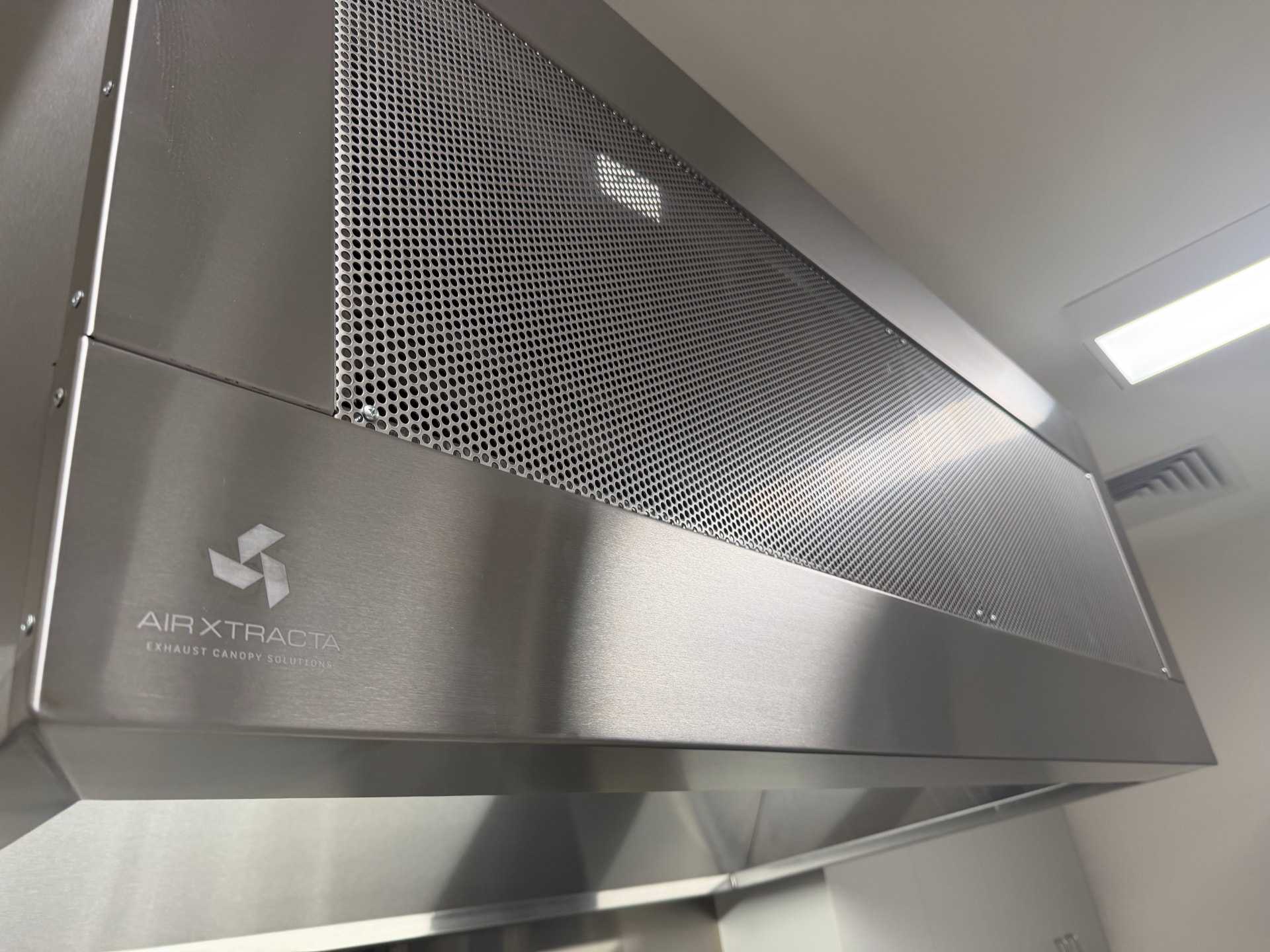 Close-up of an Airxtracta stainless canopy with perforated makeup air panel and etched brand mark